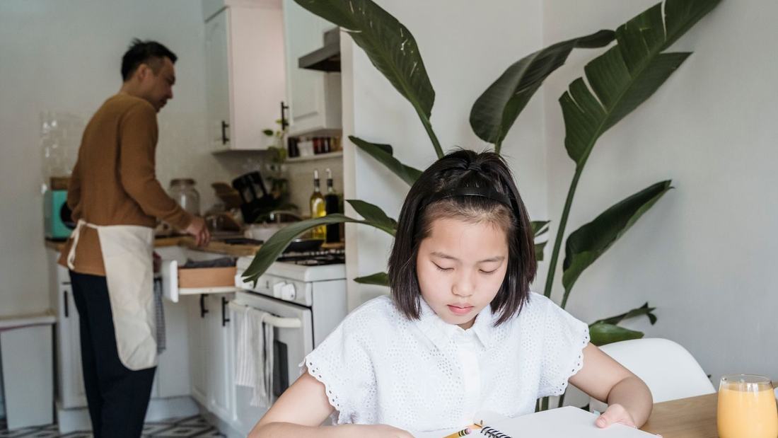 Dziewczynka odrabia lekcje przy stole, w tle tata w kuchni